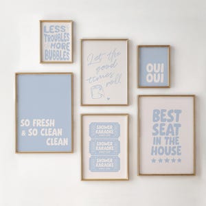 May include: Several framed art prints with gold frames and light blue and white text. The prints display phrases such as "Less Troubles & More Bubbles," "Oui Oui," and "Shower Karaoke." The art is displayed on a white wall.