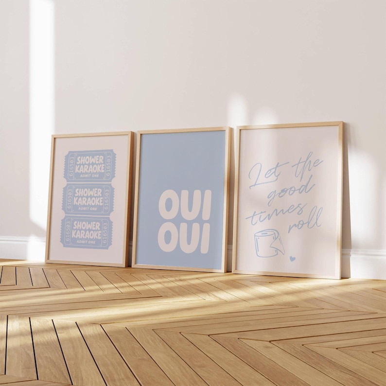 Images shows 3 funny bathroom prints with wood frames, displaying text-based designs in pastel blue & white. The artwork includes phrases such as "Oui Oui" and "Let The Good Times Roll", suitable for bathroom decor.