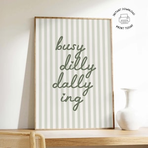 May include: A framed print with a light brown wooden frame. The print has a striped background in light green and white. The words "busy dilly dallying" are written in a cursive font in the center of the print. A white vase sits on a wooden shelf.