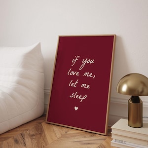 以下が含まれることがあります： 「if you love me, let me sleep」の白文字が書かれた、小さなハートが下部にある、フレーム入りのバーガンディ色のプリント。 金色のランプと本の山が右側にあります。 白いビーンバッグチェアが左側にあります。
