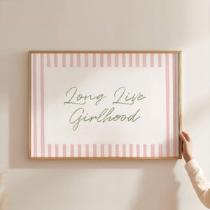 Könnte beinhalten: Ein gerahmter Druck mit dem Schriftzug "Long Live Girlhood" in Schreibschrift. Der Druck hat einen weißen Hintergrund mit rosa vertikalen Streifen an den Rändern. Der Rahmen ist aus hellem Holz.