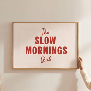 Könnte beinhalten: Ein gerahmter Druck mit den Worten "The SLOW MORNINGS Club" in roter Schrift. Der Druck hat einen cremefarbenen Hintergrund und einen Rahmen aus Naturholz. Die Hand einer Person ist sichtbar, die den Rahmen hält.