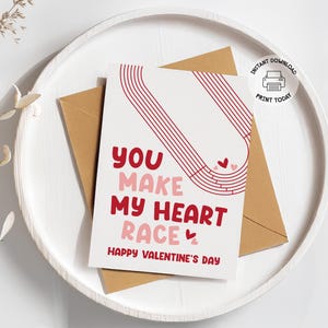 Puede incluir: Una tarjeta del Día de San Valentín con el texto "You Make My Heart Race" en rojo y rosa, junto con un gráfico de una pista de carreras. La tarjeta está en una bandeja blanca con un sobre marrón.
