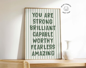 Op de afbeelding: Een ingelijste print met een lichtgroene en witte gestreepte achtergrond. De tekst luidt "YOU ARE STRONG BRILLIANT CAPABLE WORTHY FEARLESS AMAZING" in donkergroen. Een witte vaas staat op een houten oppervlak.