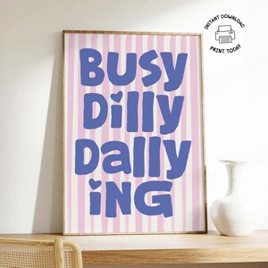 May include: A framed art print with the words "Busy Dilly Dallying" in blue, set against a pink and white striped background. The print is displayed on a wooden shelf with a white vase.