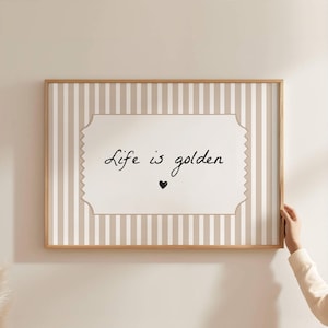 Peut inclure: Une affiche murale rectangulaire dans un cadre en bois clair avec un fond à rayures verticales beiges et blanches. Une étiquette centrale blanche aux bords festonnés porte le texte manuscrit noir Life is golden au-dessus d'un petit cœur noir.