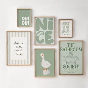 Op de afbeelding: Een verzameling ingelijste kunstprints met gouden frames. De prints tonen verschillende teksten en illustraties in groen- en wittinten, waaronder zinnen als "Oui Oui" en "Best Seat in the House". Het thema is badkamerdecoratie.