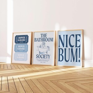May include: Three framed wall art prints. The left print shows three blue ticket stubs labeled SHOWER KARAOKE. The middle print features a bathtub illustration with the text THE BATHROOM SOCIETY. The right print displays the text NICE BUM! in blue.