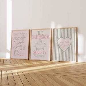 May include: Three framed art prints with light wood frames. The prints feature text and illustrations in pink, green, and white. The first print says "Guest Check Let the good times roll". The second print says "The Bathroom Society". The third print says "you look so good".