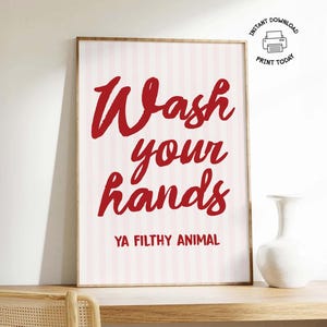 May include: A framed print with the words "Wash your hands" in red script, with "YA FILTHY ANIMAL" below. The background is pink and white striped. A white vase sits on a wooden surface.
