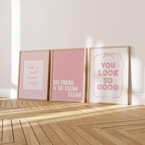 Puede incluir: Tres láminas decorativas rectangulares en marcos de madera clara. La de la izquierda tiene rayas rosas y el texto "take a seat, sweet cheeks". La central es de color rosa sólido con el texto "SO FRESH & SO CLEAN CLEAN". La de la derecha tiene el texto "YOU LOOK SO GOOD" dentro de un contorno rosa.