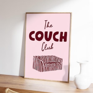 May include: A framed pink print with the words "The COUCH Club" in burgundy lettering. Below the text is a burgundy and white striped sofa illustration. The print is displayed on a wooden surface.