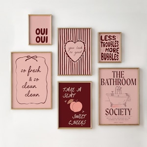 May include: A collection of framed art prints with gold frames. The prints feature various phrases and illustrations in shades of pink and burgundy, including "Oui Oui", "So Fresh & So Clean", and "The Bathroom Society".