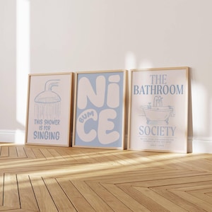 May include: Three rectangular wall art prints in light blue and white. The left print shows a shower head with the text "THIS SHOWER IS FOR SINGING". The center print features the text "NICE BUM". The right print shows a bathtub with "THE BATHROOM SOCIETY".
