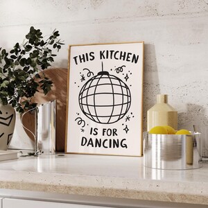 May include: A framed print with the text "THIS KITCHEN IS FOR DANCING" in black. The print features a disco ball illustration. The image is set on a white countertop with decorative items.