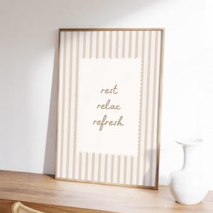 Könnte beinhalten: Ein gerahmter Druck mit beigefarbenem und weißem Streifenhintergrund und den Worten "rest, relax, refresh" in einer dekorativen Schriftart. Rechts vom Rahmen steht eine kleine weiße Vase auf einer Holzoberfläche.