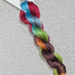 May include: A skein of hand-dyed cross stitch floss in a gradient of colors, including blue, red, green, orange, and brown. A white tag reads "Dyeing for Cross Stitch" and "Hand Dyed Fabric and Floss."