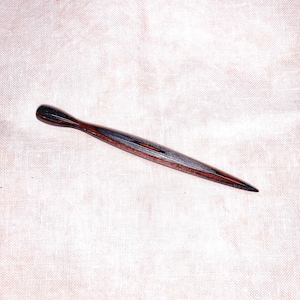 May include: A handcrafted wooden knitting needle with a tapered design. The needle features a dark, reddish-brown and black striped pattern. The tip is finely pointed, and the handle is rounded. The needle is set against a light pink fabric background.