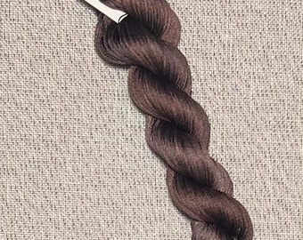 May include: A skein of hand-dyed brown cotton floss for cross stitch. The floss is twisted and has a tag that reads "Dyeing for Cross Stitch" and "Cotton". The background is a textured, neutral fabric.