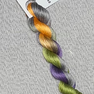 May include: A skein of hand-dyed embroidery floss in shades of purple, green, gold, and gray. The floss is labeled "Dyeing for Cross Stitch" and "Hand Dyed Fabric and Floss".