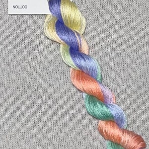 May include: A skein of hand-dyed cotton embroidery floss in a rainbow of colors, including yellow, blue, orange, green, and purple. The floss is displayed on a piece of gray fabric with the text "Dyeing for Cross Stitch Hand Dyed Fabric and Floss www.dyeing4crossstitch.com Cotton" on a white card.