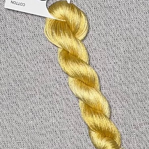 Puede incluir: Un ovillo de hilo de algodón amarillo brillante para punto de cruz, con una etiqueta que dice "Dyeing for Cross Stitch, Hand Dyed Fabric and Floss". El hilo está torcido y descansa sobre una superficie gris texturizada.