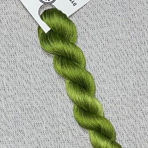 May include: A skein of olive green cotton embroidery floss, hand-dyed for cross stitch. A white tag with the text "Dyeing for Cross Stitch" and a website address is attached to the floss. The floss is on a gray textured background.