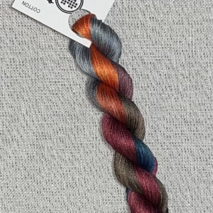 May include: A skein of hand-dyed cotton embroidery floss featuring a variegated color palette of copper, charcoal gray, deep burgundy, and navy blue. A white paper tag is attached, printed with the text DYEING FOR CROSS STITCH.
