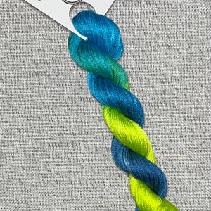 May include: A skein of hand-dyed embroidery floss in shades of blue and green. The floss is wrapped around a white card with the text "Dyeing For Cross Stitch" and "Hand Dyed Fabric and Floss" printed on it. The card also has a small image of a hoop with cross stitches.