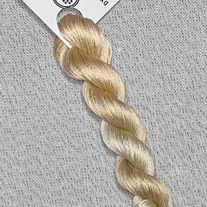 May include: A skein of light beige cotton embroidery floss, twisted and displayed against a textured gray background. A white tag attached to the floss reads "Dyeing for Cross Stitch" and "Cotton."