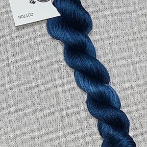 May include: A skein of dark blue cotton embroidery floss. A white tag is attached, with the text "Dyeing for Cross Stitch, Hand Dyed Fabric and Floss" and a website address. The floss is twisted and has a silky appearance.