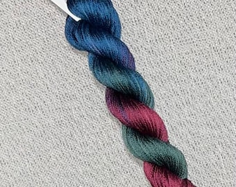 May include: A skein of hand-dyed cotton floss in shades of teal, green, and burgundy. The floss is labeled "Dyeing for Cross Stitch" and is attached to a white tag with the word "Cotton" printed on it.