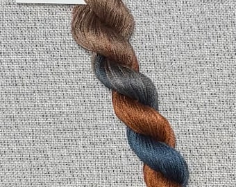 May include: A twisted bundle of cotton embroidery floss in shades of brown, gray, and blue. A white tag with the text "Dyeing for Cross Stitch" and a website address is visible. The floss is on a gray textured fabric.