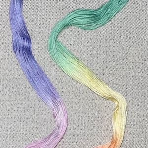 May include: A rainbow-coloured embroidery floss skein in shades of blue, green, yellow, pink, and orange. The floss is arranged in a curved shape on a grey fabric background.