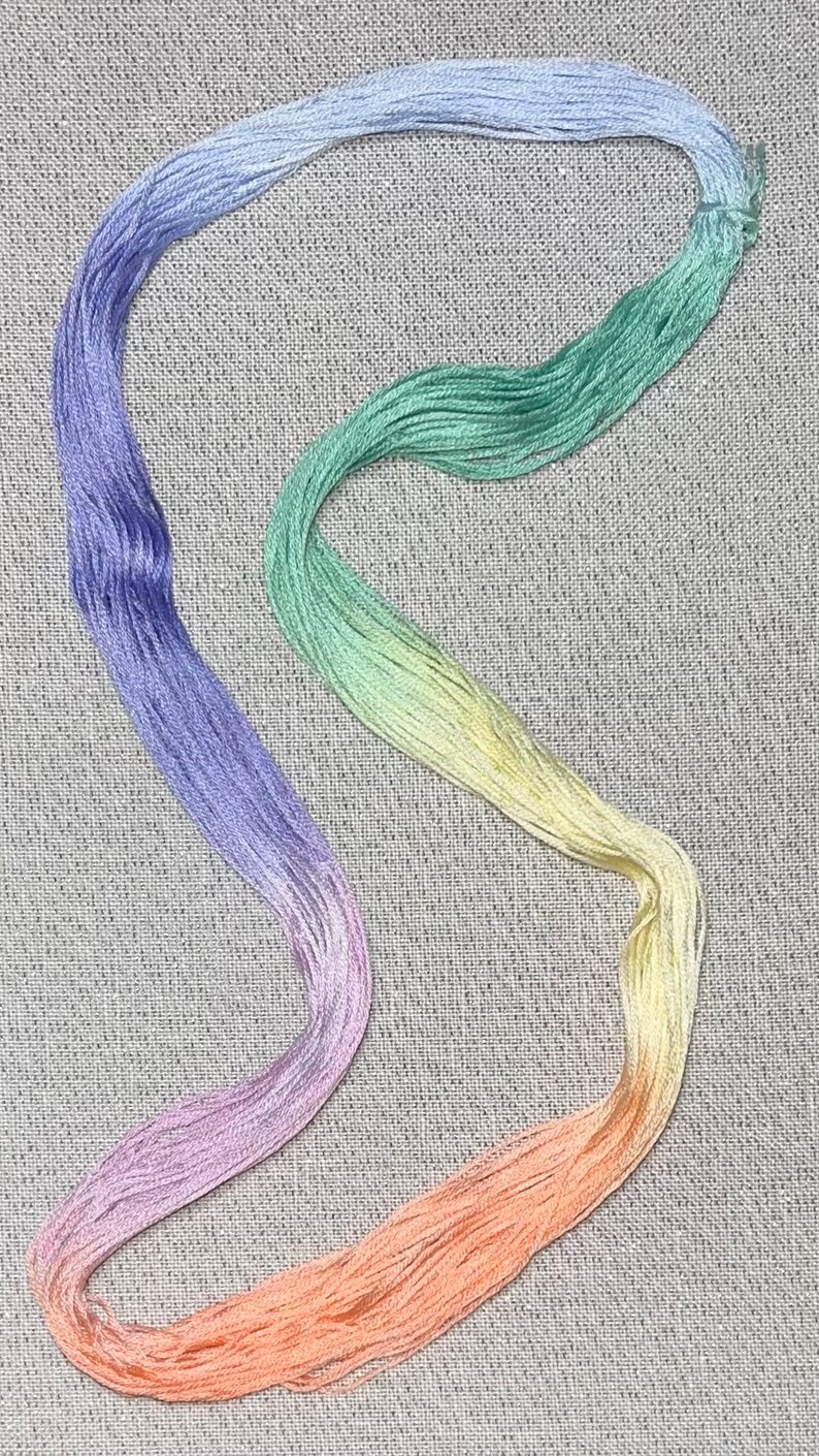 May include: A rainbow-coloured embroidery floss skein in shades of blue, green, yellow, pink, and orange. The floss is arranged in a curved shape on a grey fabric background.
