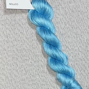 May include: A skein of light blue cotton embroidery floss for cross stitch. The floss is hand-dyed and has a twisted appearance. A white tag with the text "Dyeing for Cross Stitch" is attached.
