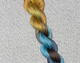 May include: A skein of hand-dyed cotton floss in shades of gold, blue, and brown. The floss is twisted and has a tag that reads "Dyeing for Cross Stitch" and "Hand Dyed Fabric and Floss."