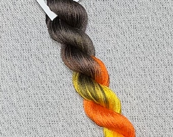 May include: A skein of hand-dyed embroidery floss in shades of brown, yellow, and orange. The floss is labeled "Dyeing for Cross Stitch" and "Hand Dyed Fabric and Floss".