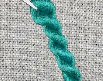 May include: A skein of teal blue hand-dyed cotton embroidery floss for cross stitch. The floss is twisted and coiled on a white card with the text "Dyeing For Cross Stitch" and "Hand Dyed Fabric and Floss" printed on it. The card also has a website address printed on it: www.dyeingforcrossstitch.com.