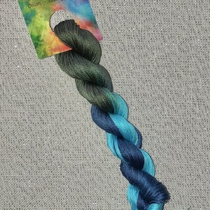 Puede incluir: Un ovillo de hilo de bordar de algodón teñido a mano en tonos de verde, azul y turquesa. El hilo está etiquetado "Dyeing for Cross Stitch - Cotton - hand dyed fibers".