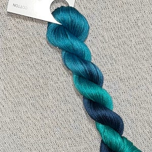 May include: A skein of hand-dyed cotton floss in shades of teal, turquoise, and navy blue. The floss is labeled "Dyeing for Cross Stitch" and is displayed on a gray textured surface. The floss is twisted into a rope-like shape.
