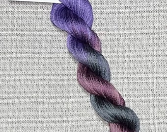 May include: A skein of hand-dyed cotton floss for cross stitch in shades of purple, gray, and burgundy. The floss is twisted and has a tag that reads "Dyeing for Cross Stitch". The background is a light gray fabric.