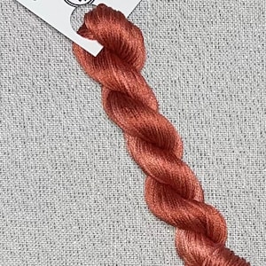 May include: A skein of hand-dyed cotton embroidery floss in a warm terracotta color. The floss is labeled "Dyeing For Cross Stitch" and "Hand Dyed Fabric and Floss" with the website address "www.dyeingforcrossstitch.com".
