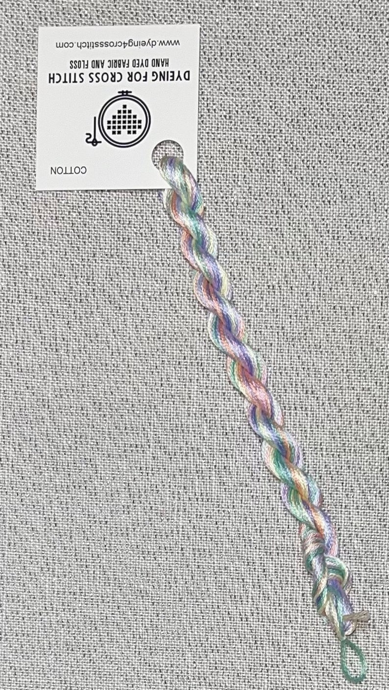 May include: A skein of hand-dyed cotton embroidery floss in shades of green, pink, and purple. The floss is wrapped around a white card with the text "Dyeing For Cross Stitch" and "Hand Dyed Fabric and Floss" and the website address "www.dyeing4crossstitch.com".