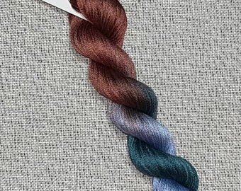 May include: A skein of hand-dyed embroidery floss in shades of brown, blue, and green. The floss is arranged in a spiral shape and is labeled "Dyeing For Cross Stitch" and "Hand Dyed Fabric and Floss".