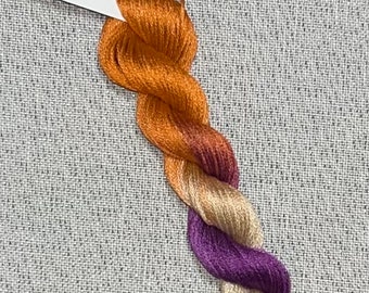 May include: A skein of hand-dyed embroidery floss in shades of orange, yellow, and purple. The floss is labeled "Dyeing For Cross Stitch" and "Cotton".