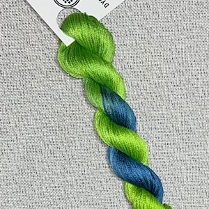 Puede incluir: Madeja retorcida de hilo de punto de cruz teñido a mano en tonos verdes y azules. Una etiqueta blanca con el texto "Dyeing for Cross Stitch" y un logotipo está adjunta. El hilo es de algodón.