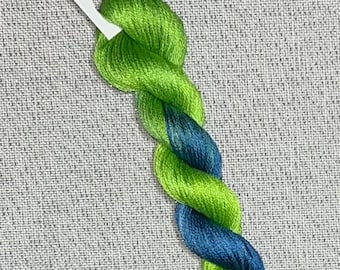 May include: A twisted skein of hand-dyed cross stitch floss in shades of green and blue. A white tag with the text "Dyeing for Cross Stitch" and a logo is attached to the floss. The floss is made of cotton.