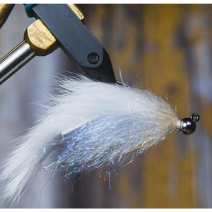 May include: Close-up of a white and silver fishing fly, held in a vise. The fly has a fluffy, feathery appearance with a silver, iridescent underlayer. A small, dark bead is at the head of the fly.