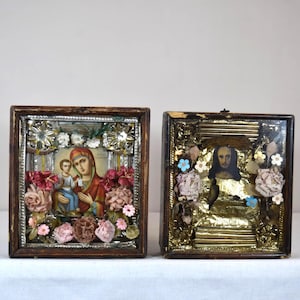 Puede incluir: Dos iconos religiosos enmarcados. Uno presenta una representación de María y Jesús, rodeados de flores rosas y rojas. El otro icono tiene un fondo dorado y un retrato de un hombre, también con detalles florales. Ambos están en marcos de madera.